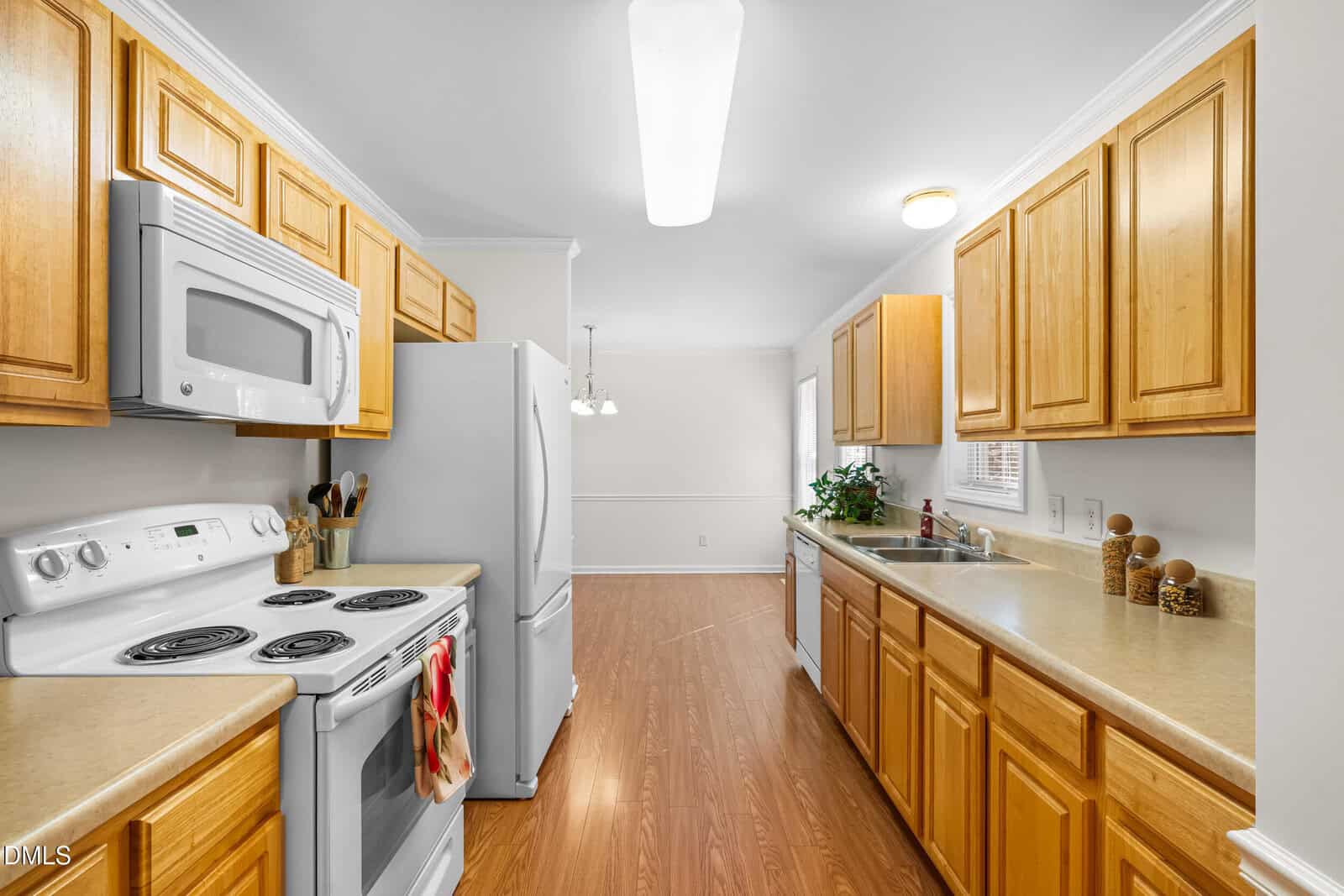 Bright kitchen with light wood cabinets, white appliances, laminate countertops, and wood flooring. A window above the sink lets in natural light. Dining area is visible in the background with a hanging light fixture.