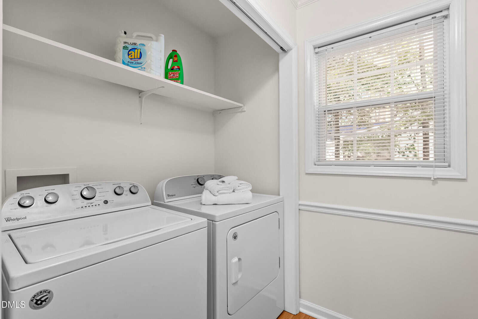A laundry area with a white Whirlpool washing machine and dryer, folded white towels on the dryer, detergent and cleaning products on a shelf above, and a window with blinds letting in natural light.