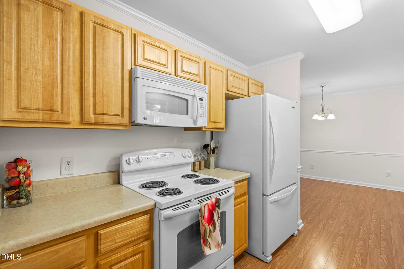 A kitchen with light wood cabinets, a white stove, microwave, and refrigerator, beige countertops, and a jar of colorful peppers. The space leads to a dining area with a hanging light fixture and wood flooring.
