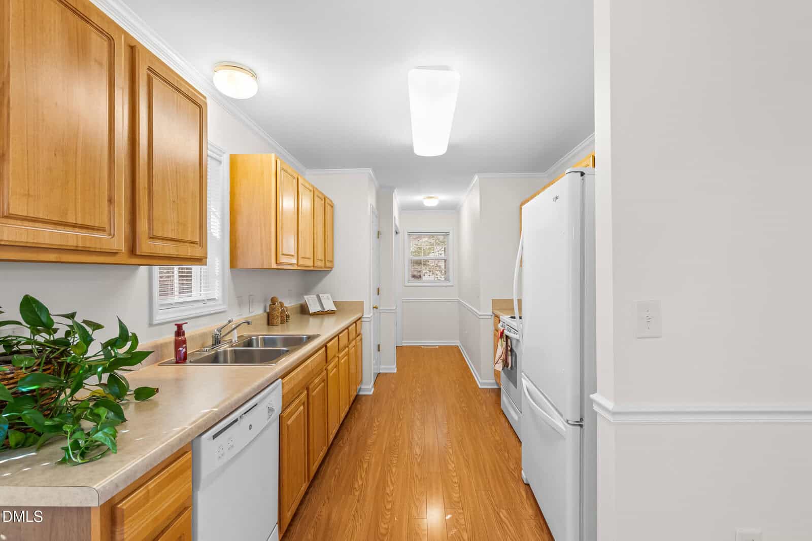 A galley kitchen with wood cabinets, laminate countertops, white appliances, and wood-look flooring. A small window is above the sink, and a green plant sits on the counter. The kitchen leads to a door at the far end.