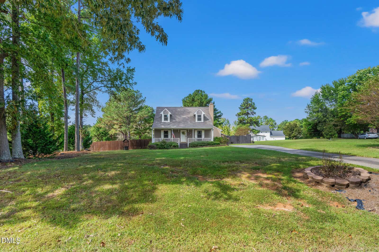 A two-story house with a front porch sits on a large grassy yard with scattered trees. A curved driveway leads to the house, and there is a fire pit area in the foreground. The sky is clear with a few clouds.