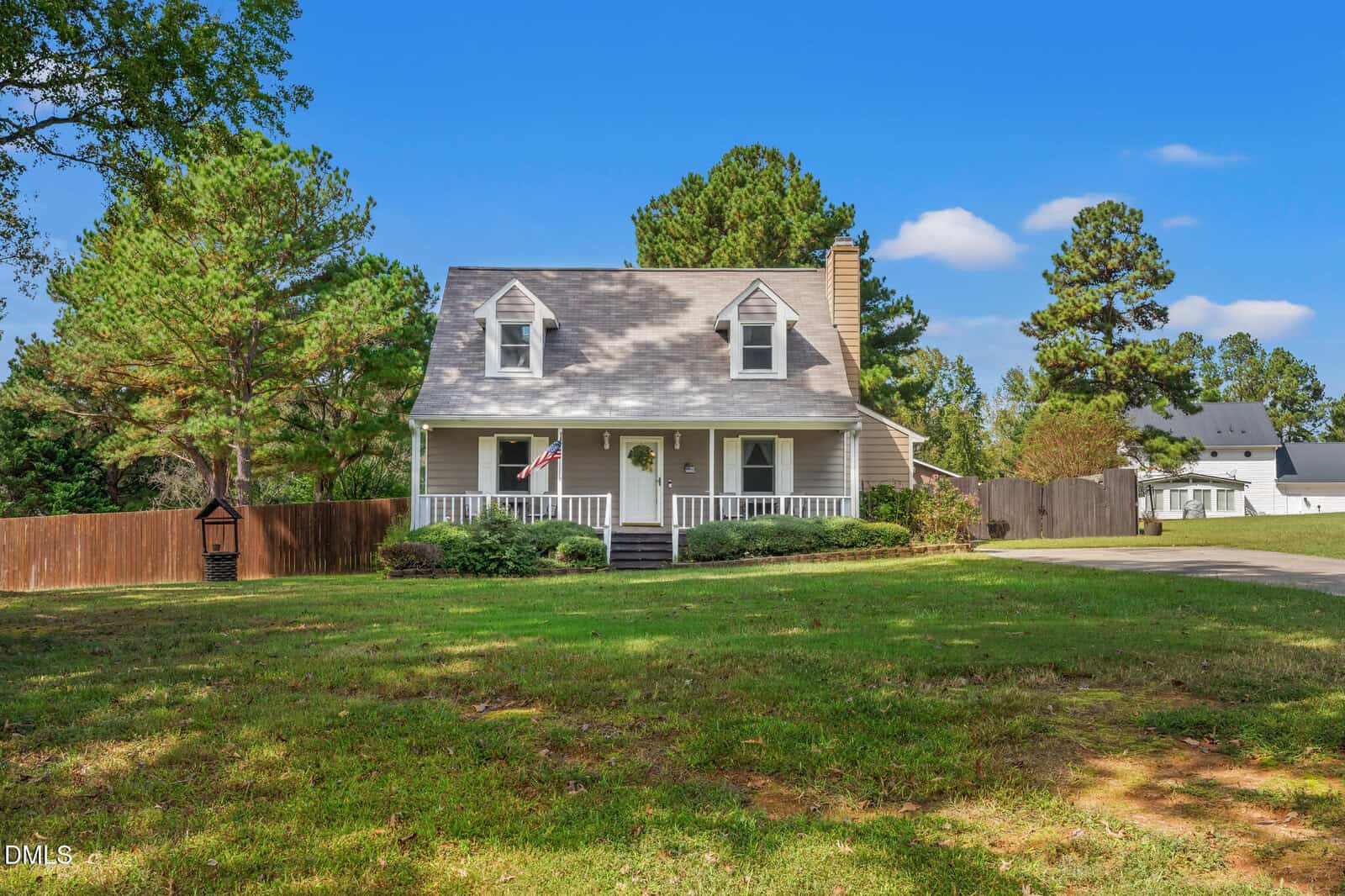 A two-story house with gray siding, a covered front porch, and dormer windows, set on a spacious lawn with trees in the background and a paved driveway to the right. An American flag hangs by the front door.