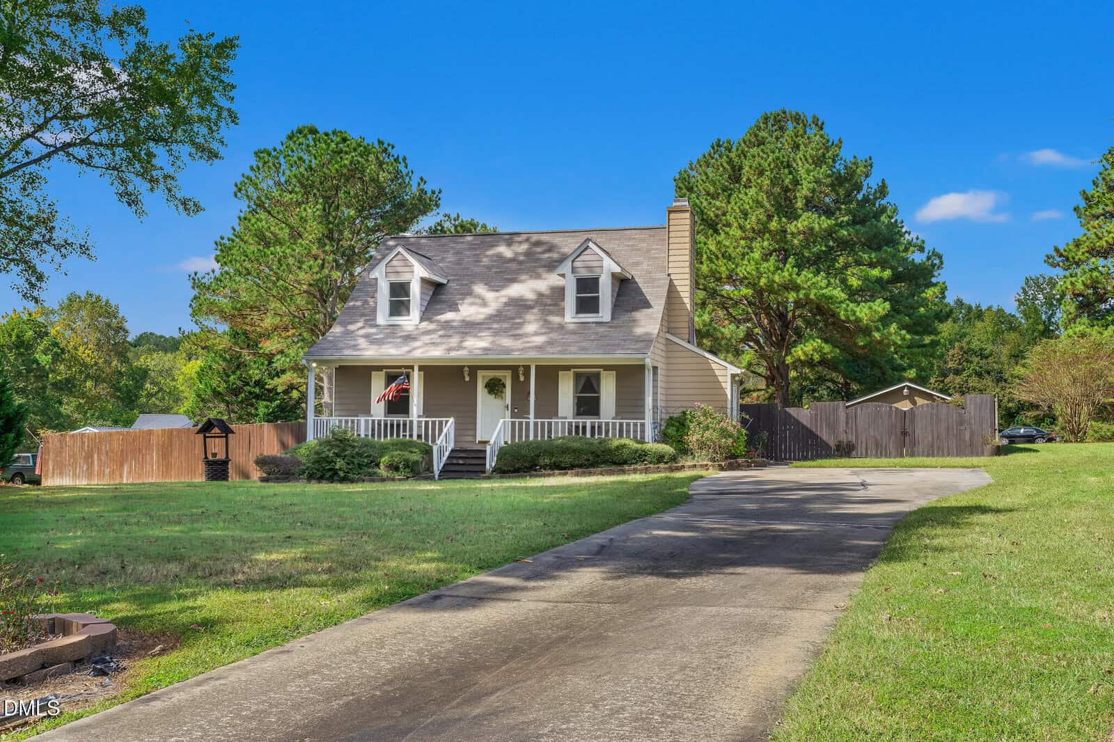 A beige two-story house with a front porch, gable roof, and dormer windows sits at the end of a curved driveway, surrounded by a lawn, trees, and a wooden fence under a clear blue sky.