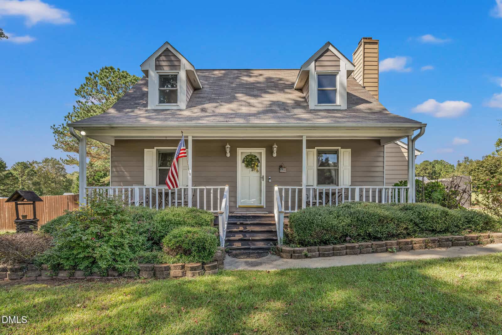 A beige two-story house with a covered front porch, two dormer windows, an American flag, and surrounding shrubs, pictured on a sunny day with a blue sky.