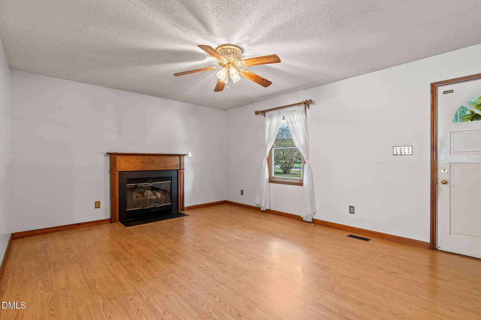 A living room with light wood flooring, a white ceiling with a ceiling fan, a fireplace with a wood mantel, a window with white curtains, and a white door with a gold handle.
