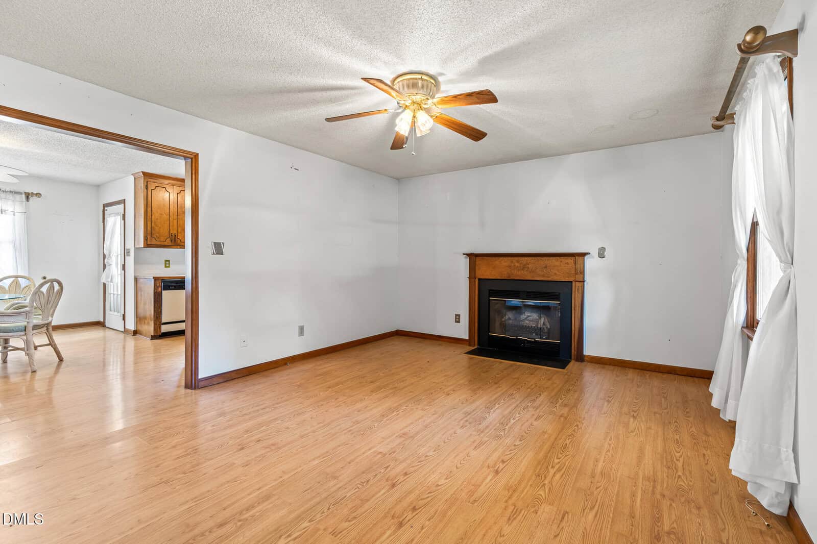 A living room with light wood flooring, white walls, a ceiling fan with lights, a wooden fireplace, and windows with sheer white curtains. An adjacent dining area and kitchen are visible through an open doorway.