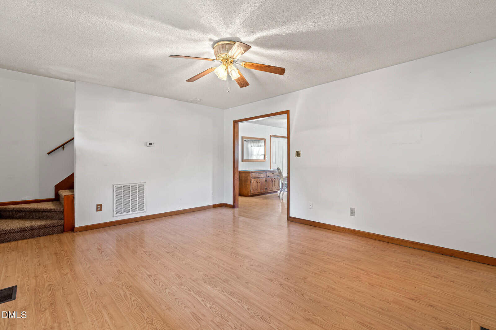 A spacious, empty living room with light-colored walls, wood laminate flooring, a ceiling fan with lights, and an open doorway leading to a dining area. Carpeted stairs are visible on the left side of the image.