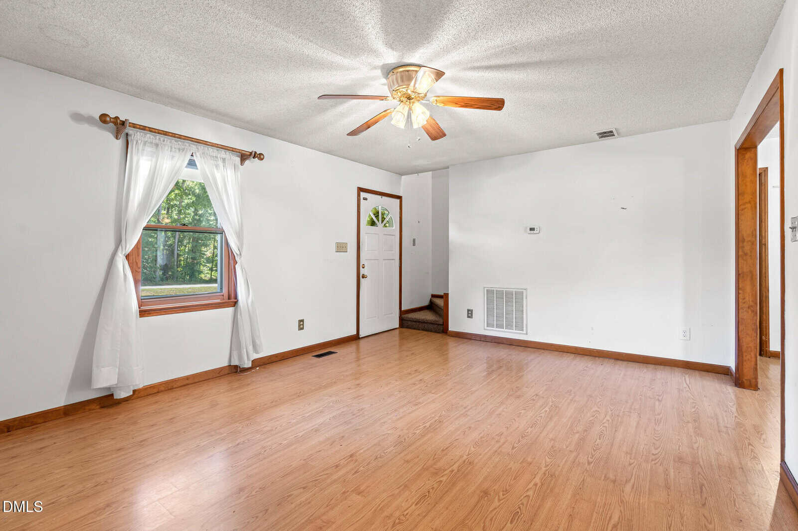 Bright, empty living room with wood floors, white walls, a ceiling fan, window with white curtains, and a white front door. An open doorway and stairs lead to other areas of the house.