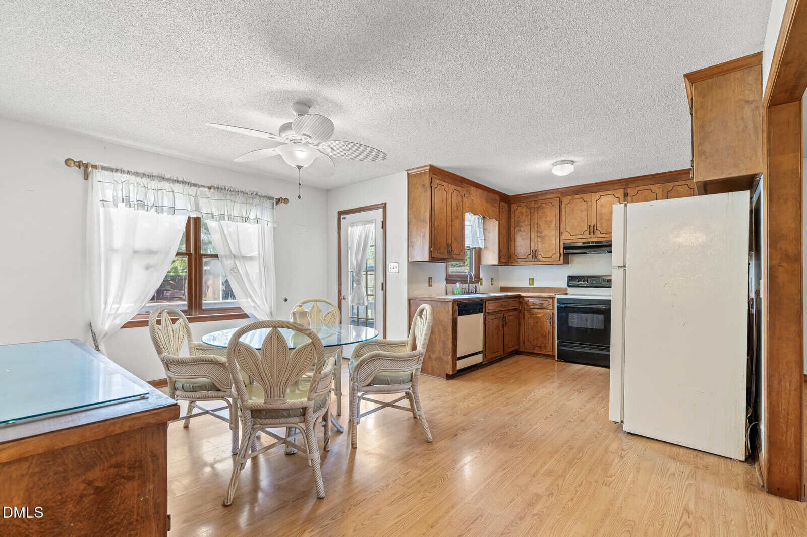 Bright kitchen with wood cabinets, white appliances, and a round glass dining table with four chairs. Large window and glass door let in natural light. Light wood flooring throughout the room.