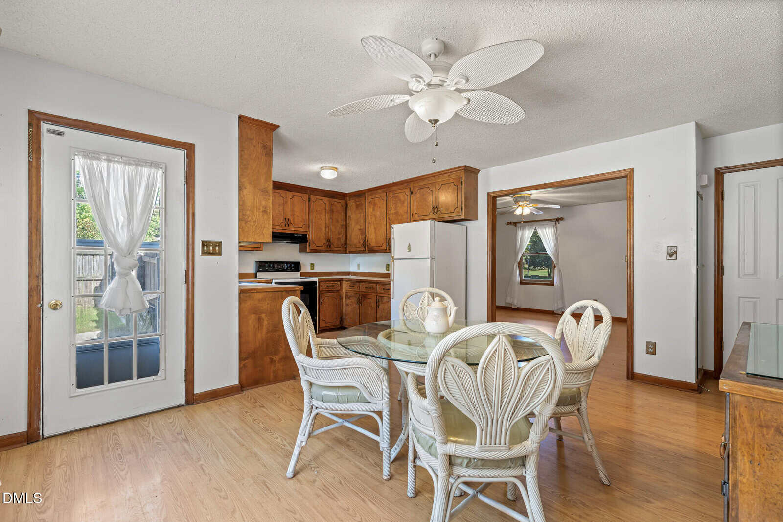 A cozy kitchen with wooden cabinets, a white fridge, and a glass dining table with four white wicker chairs. A ceiling fan is above the table, and a door with a sheer curtain leads outside.