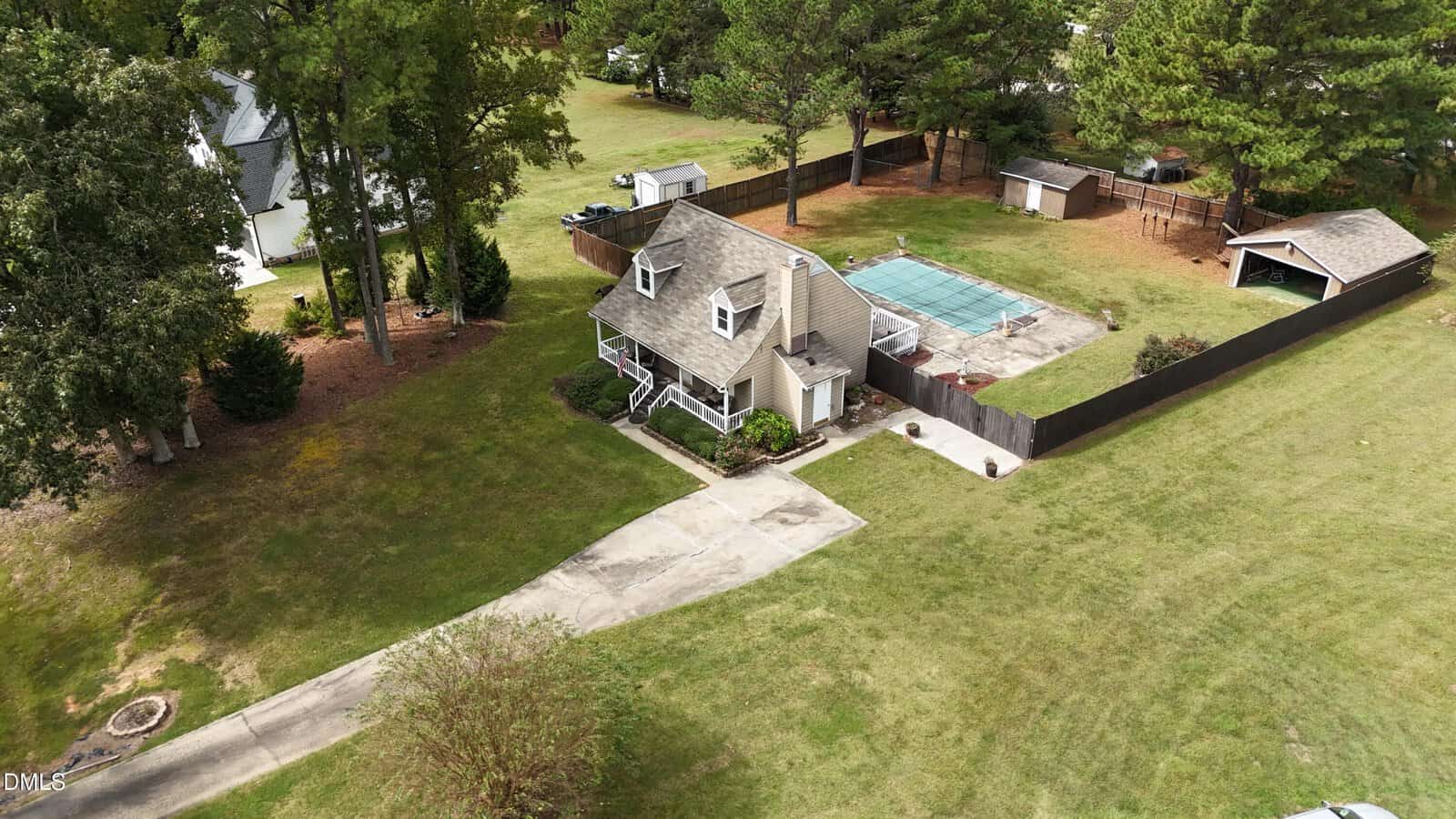 A two-story house with a fenced backyard, a covered pool, a shed, and a driveway, surrounded by grass and trees in a suburban neighborhood.
