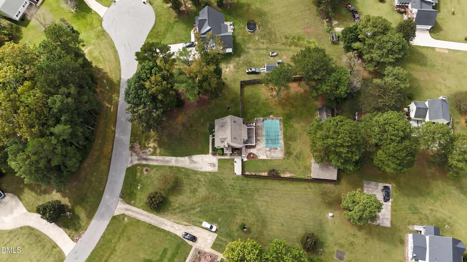 Aerial view of a suburban home with a fenced backyard, patio, and pool. The property is surrounded by trees and lawns, with neighboring houses and curved roads visible in a residential neighborhood.