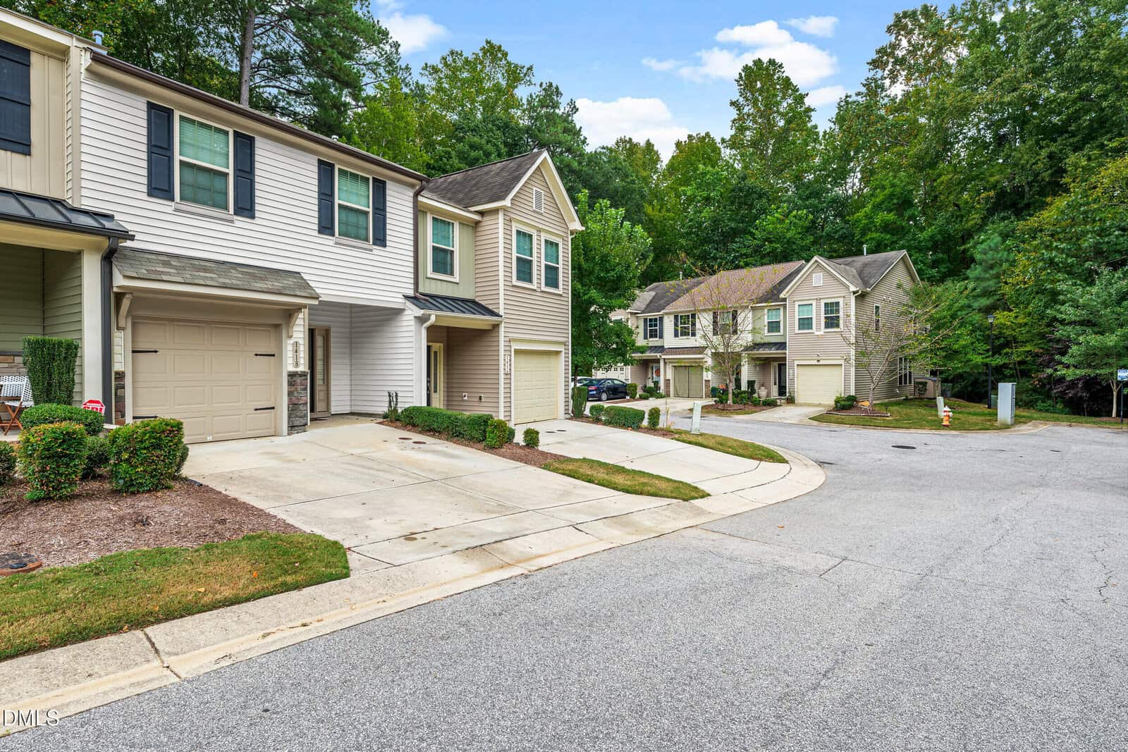 A row of modern two-story townhouses with garages sits along a curved suburban street bordered by green trees and manicured lawns under a partly cloudy sky.
