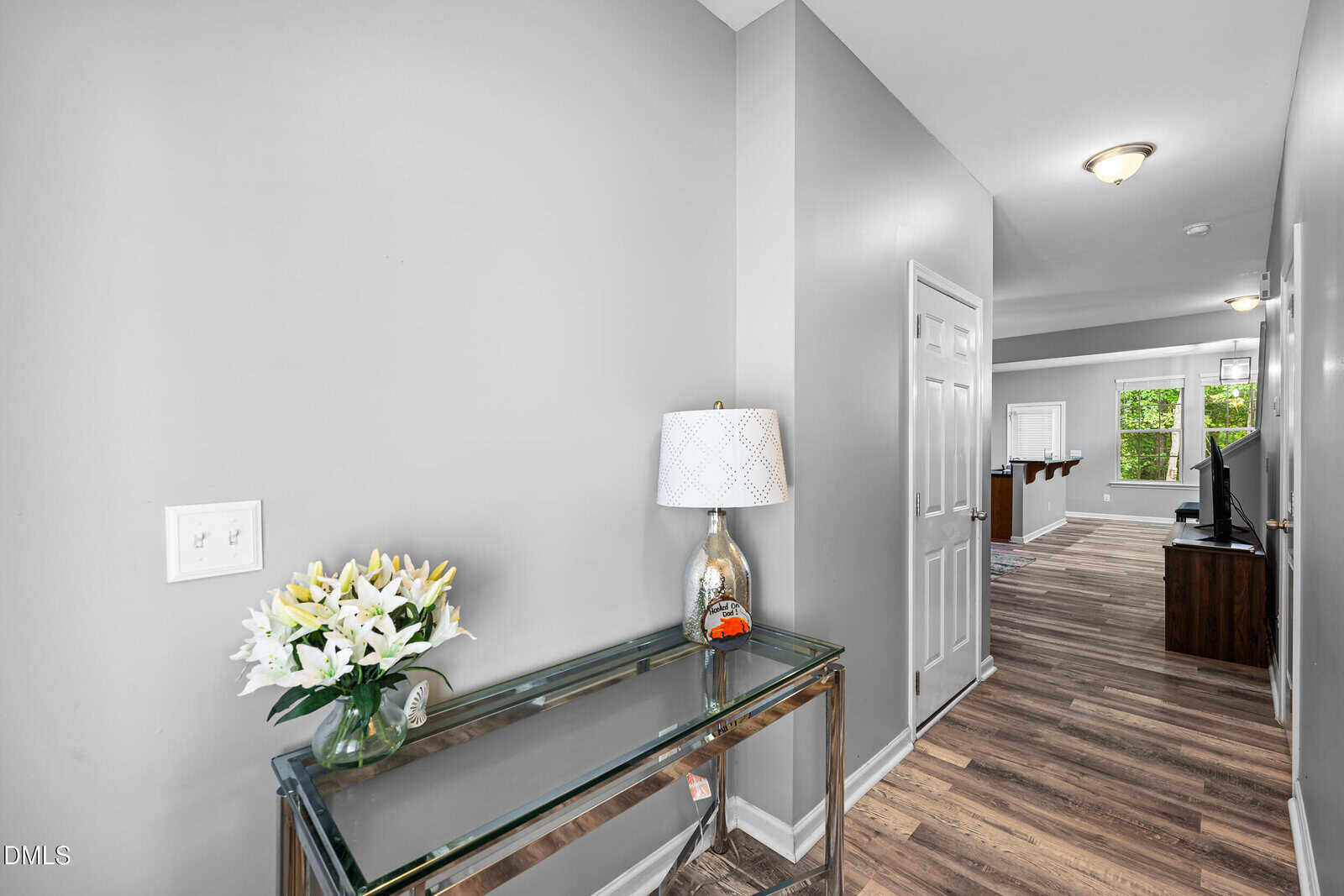A hallway with gray walls, wood-style flooring, a glass console table holding a white lamp and vase of lilies, leading to a living area with windows and a television.