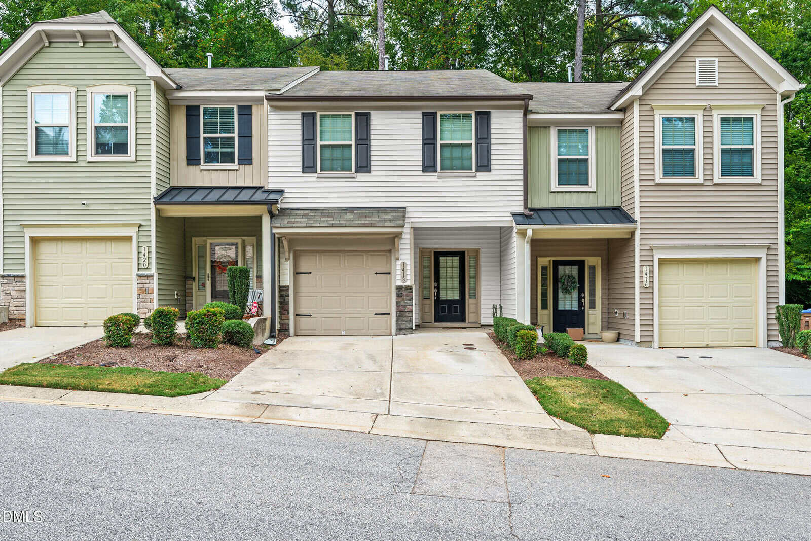 Three attached two-story townhomes at 1418 Chatuga Way, Wake Forest, feature garages, driveways, and small front porches. The exterior highlights neutral siding colors, trimmed shrubs, and a sloped street in the foreground with a wooded area behind.