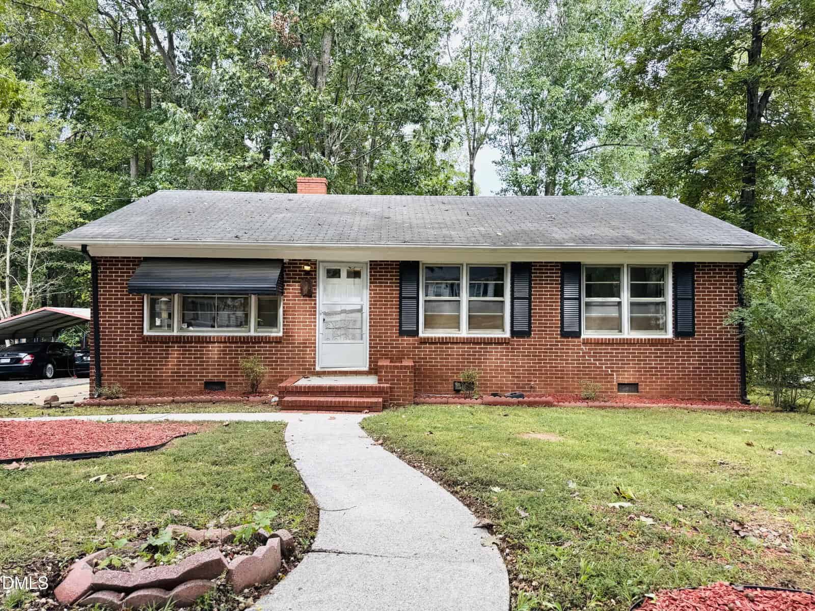Charming single-story brick house at 906 Abbott Street, Henderson, featuring a gray shingle roof, black shutters, and a white front door. A curved concrete walkway leads to the entrance, with minimal landscaping and a carport partially visible on the left.