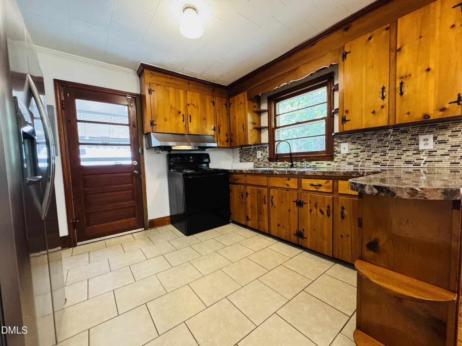 A kitchen with wooden cabinets, a black stove and range hood, mosaic tile backsplash, tile flooring, a stainless steel refrigerator, a window above the sink, and a wooden door with glass panels.