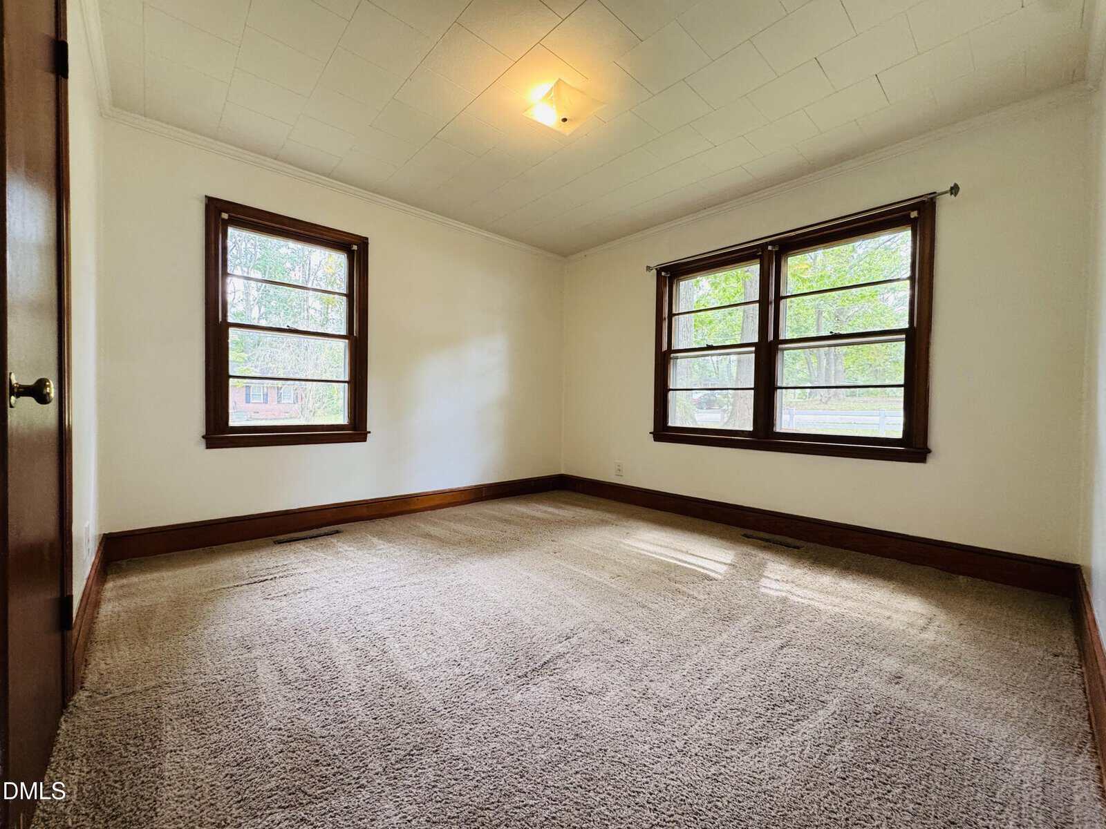 Empty bedroom with beige carpet, white walls, brown wood trim, two large windows on adjacent walls, and a ceiling light. Natural light brightens the room through the windows.