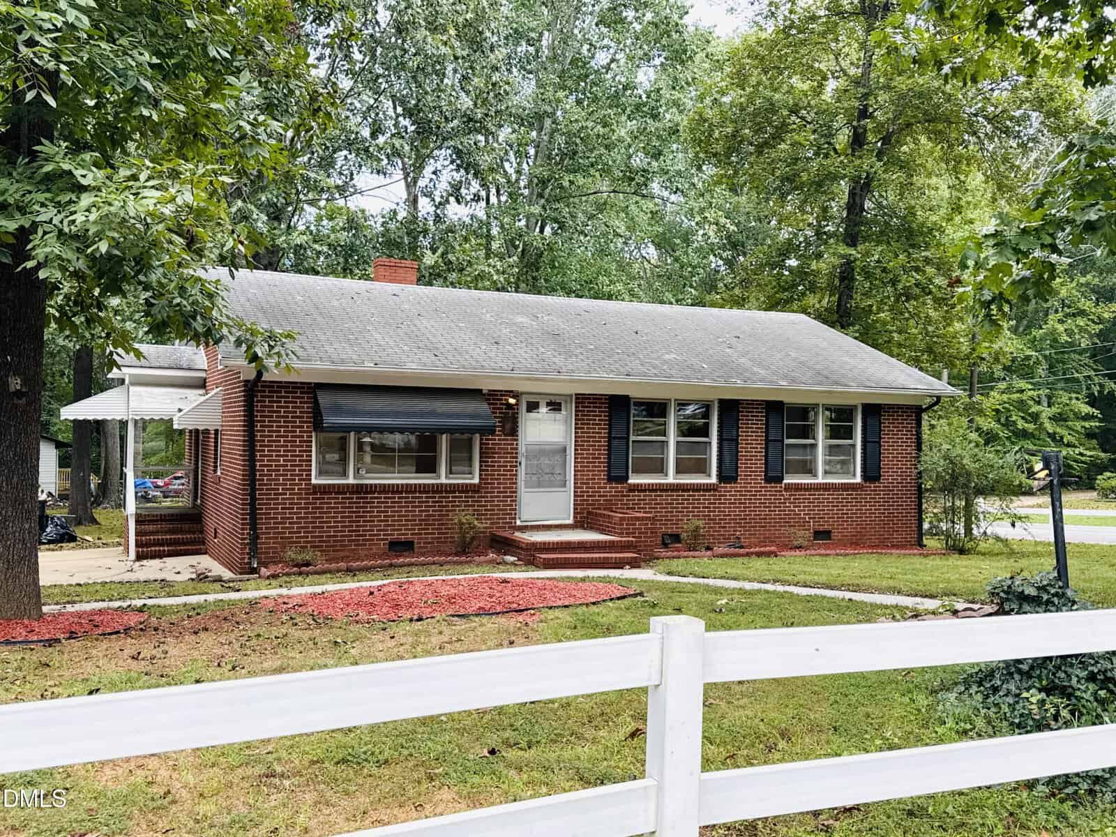 A single-story red brick house with a gray roof, white trim around the windows and door, and a covered side entrance. The yard features grass, red mulch, and a white wooden fence in the foreground.