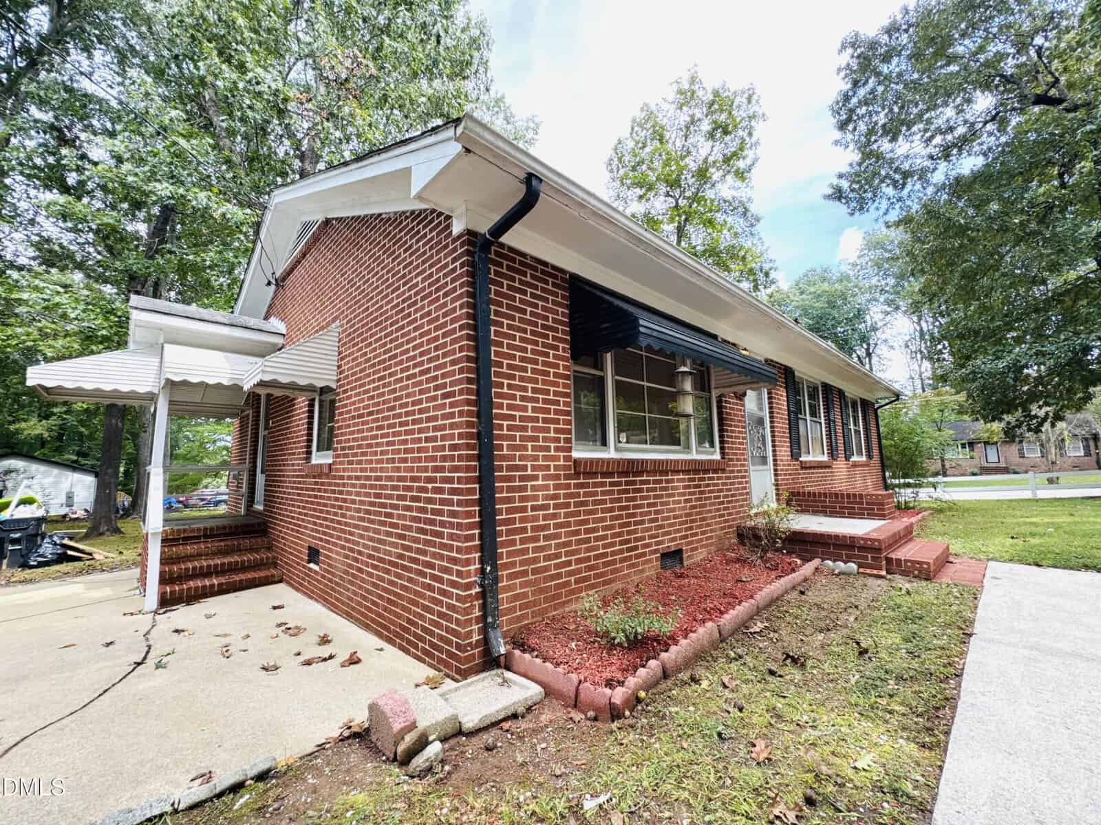 A one-story red brick house with white trim, black shutters, and a white awning over the side entrance. The yard has red mulch, small shrubs, and is surrounded by mature trees. A concrete walkway leads to the front steps.