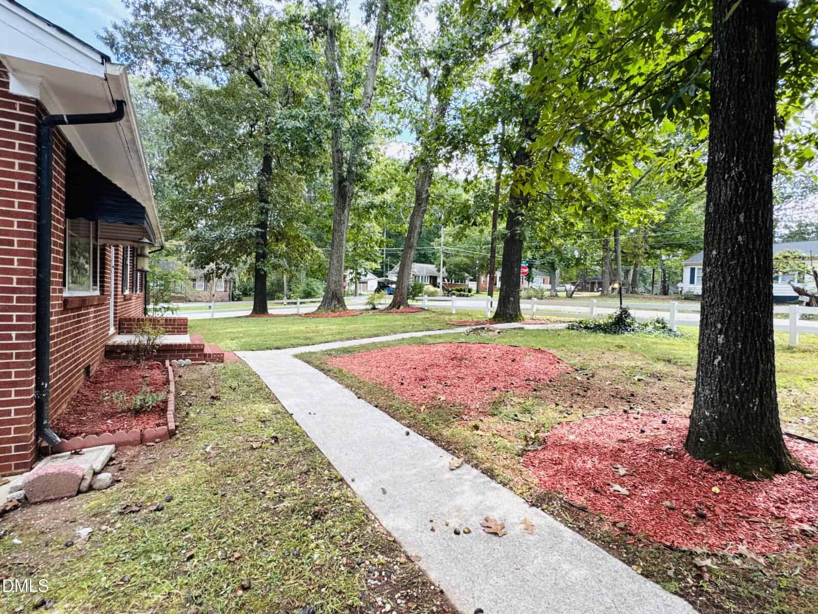 A concrete walkway leads from a red brick house to the sidewalk. The lawn features several large trees and mulched garden beds with red wood chips. Residential houses and a street are visible in the background.