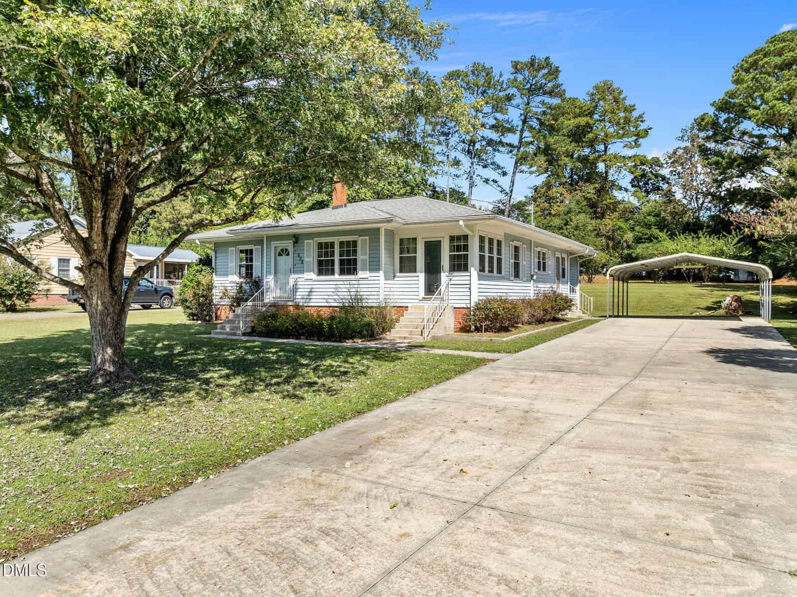 Single-story white house with a front porch, brick steps, and a large driveway leading to a metal carport. The yard has scattered trees and shrubs, with grass covering most of the ground.