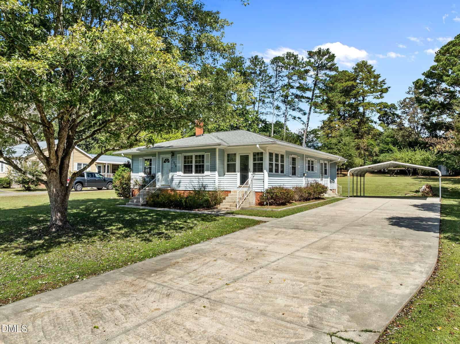 Single-story white house with a wide front porch and gray roof, set back from the street. A long concrete driveway leads to a metal carport, surrounded by green lawn, trees, and bushes under a clear blue sky.