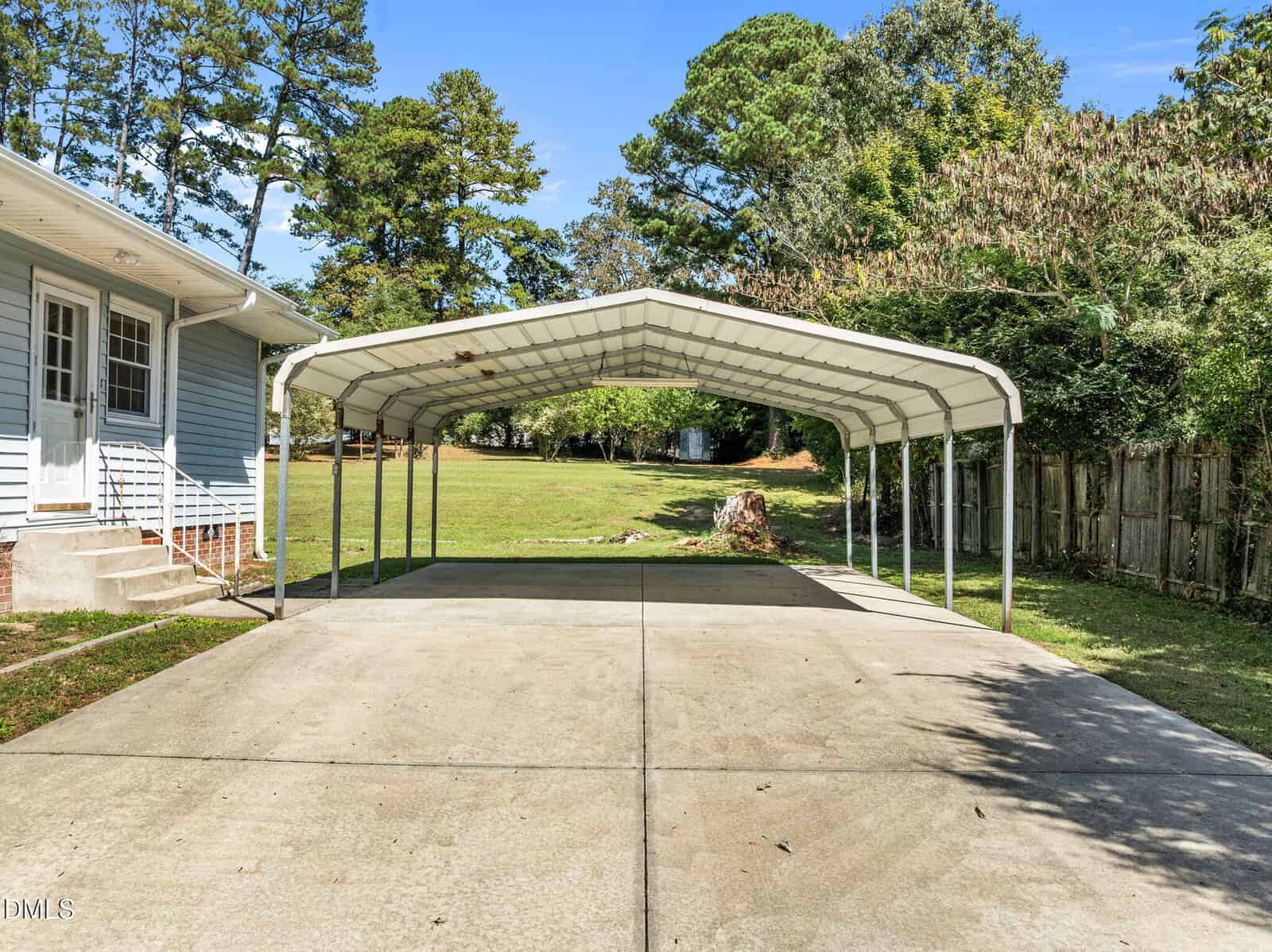 A metal carport with a peaked roof stands on a concrete driveway next to a blue house. The area is surrounded by trees and a wooden fence, with a grassy yard in the background under a clear sky.