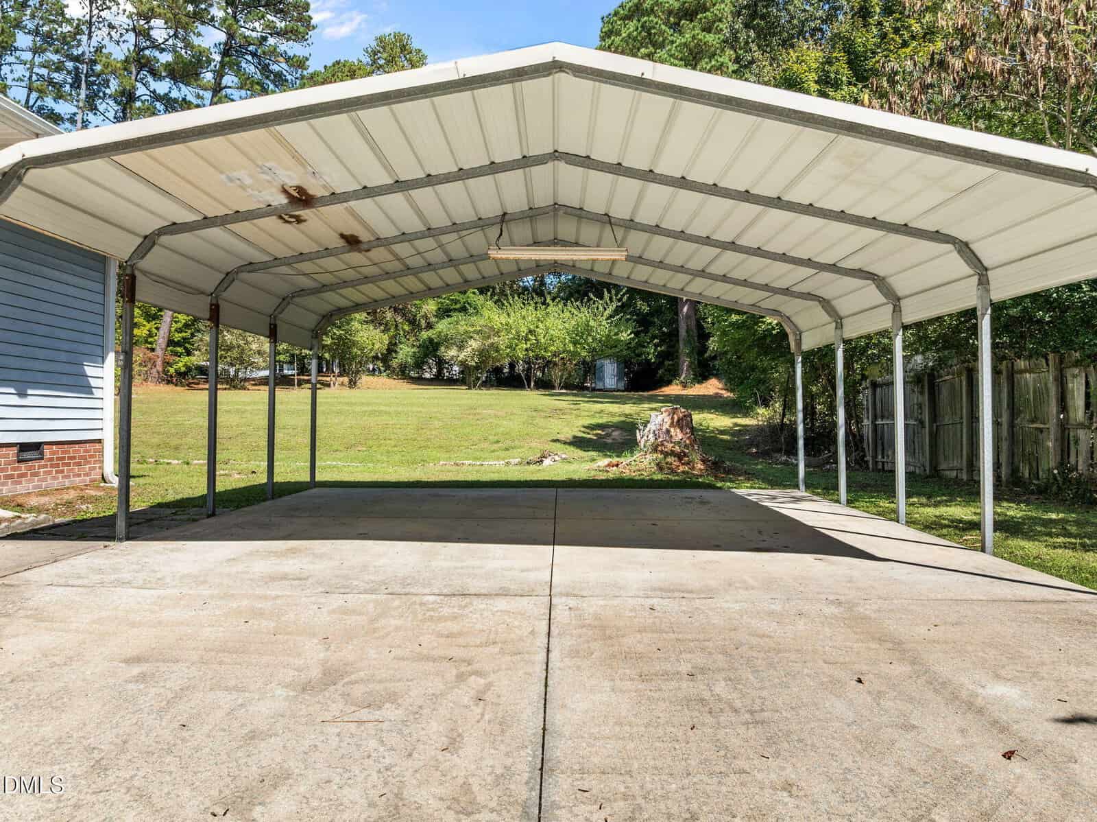A metal carport with a sloped roof stands over a concrete driveway beside a blue house. The area behind the carport is grassy with trees, a stump, and a wooden fence on the right.