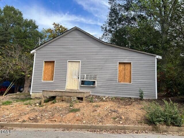 A small gray house at 505 Harriett Street in Henderson has boarded-up windows and a closed door, sitting on bare, uneven ground. An air conditioning unit is installed below one window, with trees and grass visible in the background.