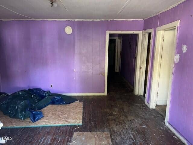 Room with purple-painted walls, visible scuff marks, and worn, uncovered wood flooring. Piles of plastic tarp and plywood are on the floor. Three doorways and a dark hallway are on the right side of the image.