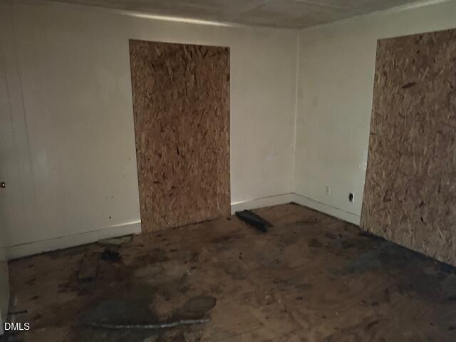 A dimly lit, empty room with boarded-up windows and walls, stained and damaged plywood floor, and plain white walls. No furniture or decor is present.
