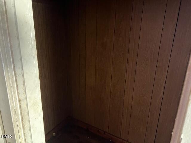 A small, empty closet or storage space with wood-paneled walls and a bare floor. The area is dimly lit, and the entrance is partially visible on the left side of the image.