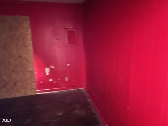 A small room with bright red walls, some scuff marks, a wooden board covering part of a doorway, a dark floor, and a red metal box mounted on the wall.
