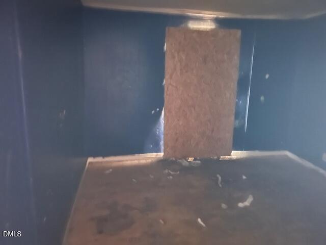 A dimly lit, empty room or box with dark walls and a wooden or particle board piece leaning against the back wall. Some debris or small scattered pieces are visible on the floor.