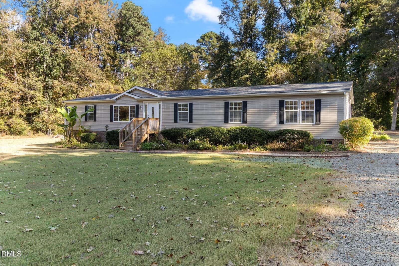 Single-story gray house with white trim and black shutters, front porch with steps, surrounded by shrubs and trees, set on a grassy lawn with a gravel driveway under a clear sky.