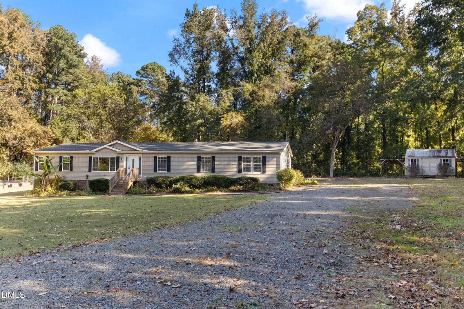 A single-story beige manufactured home with a small front porch sits on a spacious lot with a gravel driveway, surrounded by trees and a wooden shed in the background. The yard is mostly grass with some patches of dirt.