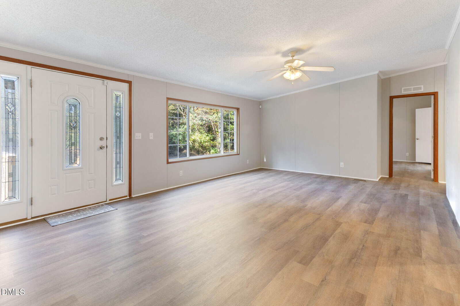 Spacious empty living room with light wood flooring, large front window, ceiling fan with light, and a white front door with glass panels. Neutral-colored walls and natural light fill the room.