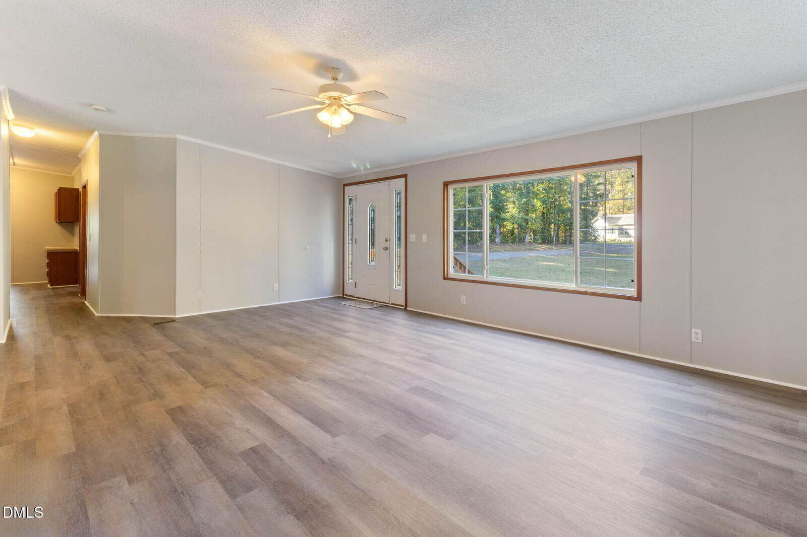 A spacious, empty living room with light wood flooring, beige walls, a ceiling fan with lights, a large front window, and a glass front door. Sunlight streams through the window, illuminating the room.