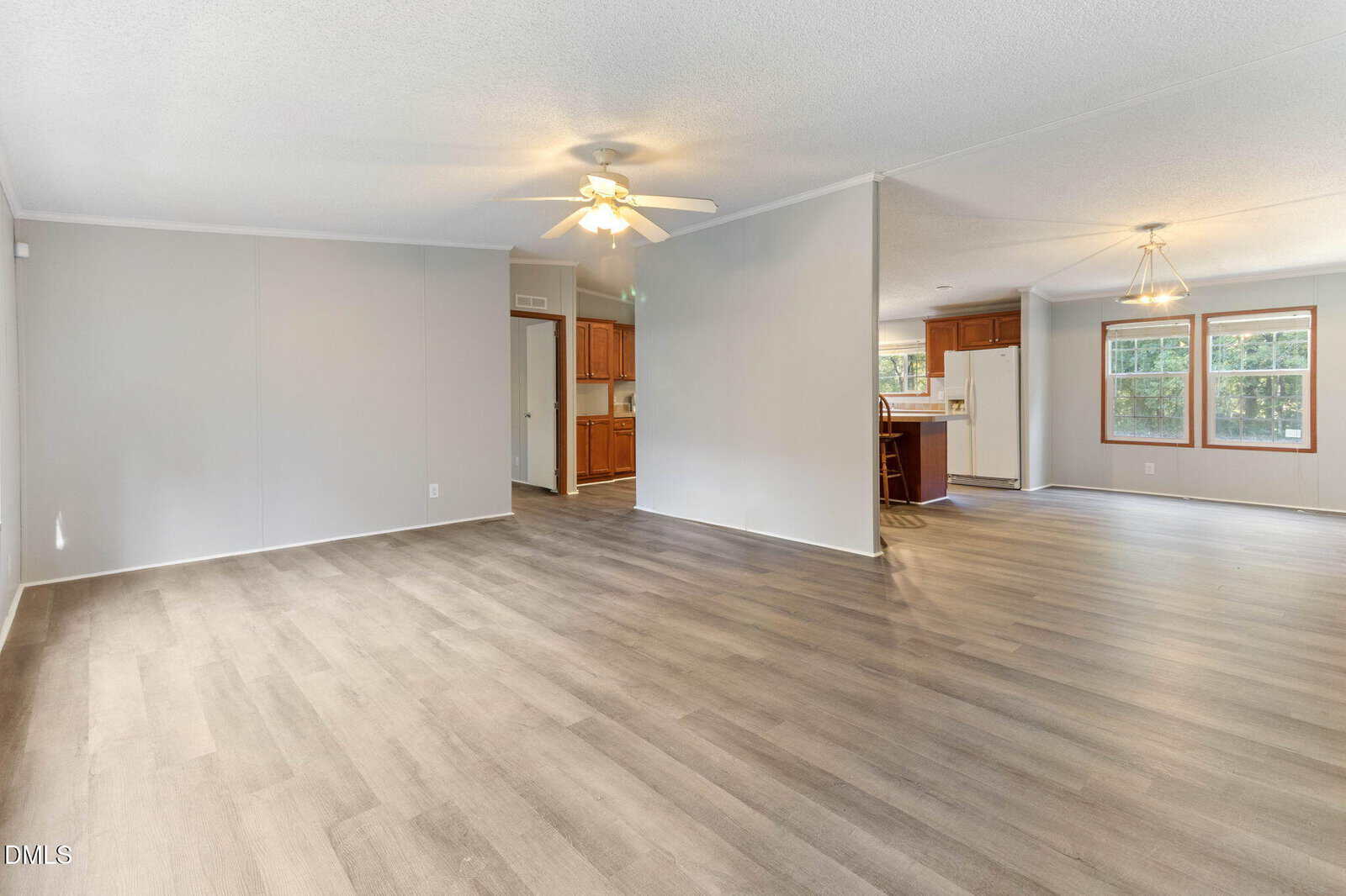 Spacious, empty open-concept living and dining area with light wood flooring, white walls, ceiling fans, and large windows. A kitchen with wood cabinets and white appliances is visible in the background.