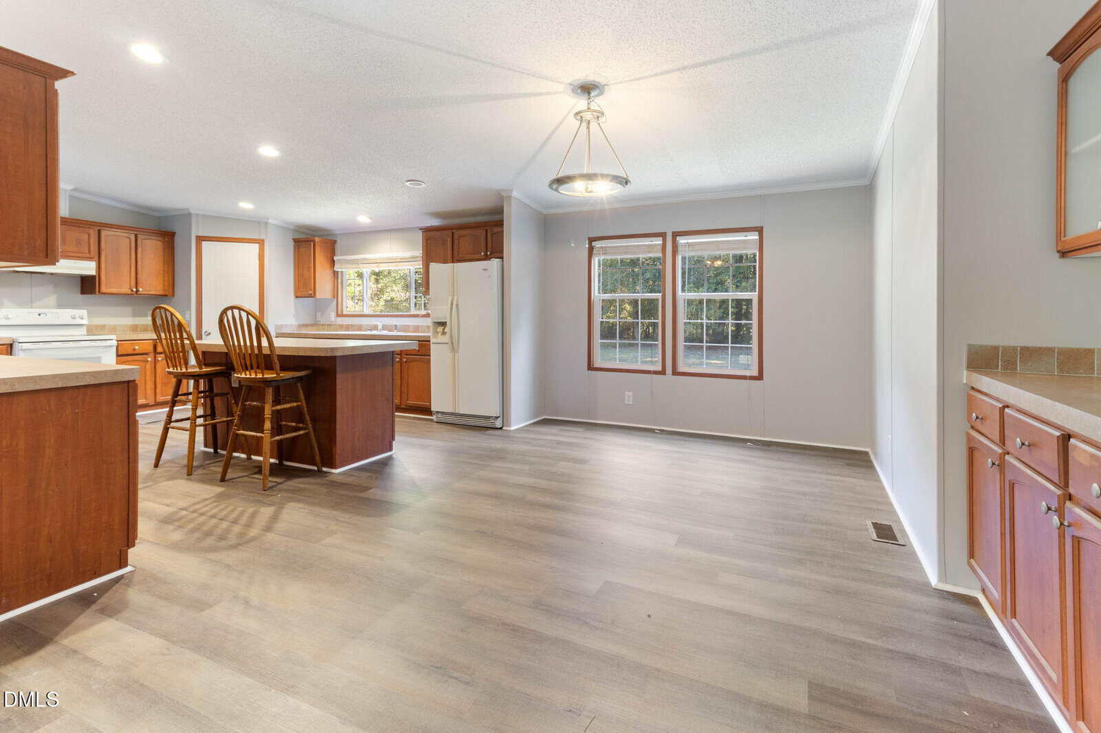 Spacious kitchen and dining area with wood cabinets, center island with two wooden chairs, white appliances, large windows, and light wood flooring. Walls are painted light gray with white trim.