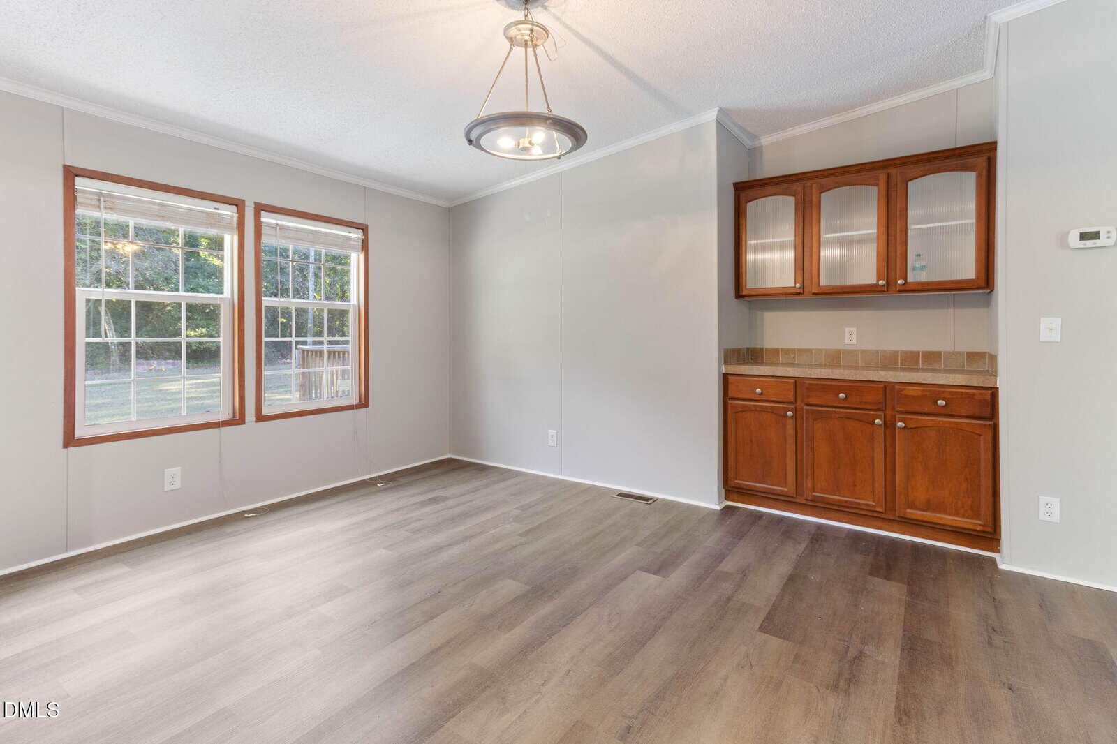 A room with light gray walls, two large windows, wood-style flooring, a ceiling light fixture, and a wooden built-in cabinet with glass doors and drawers beneath a beige tile countertop.