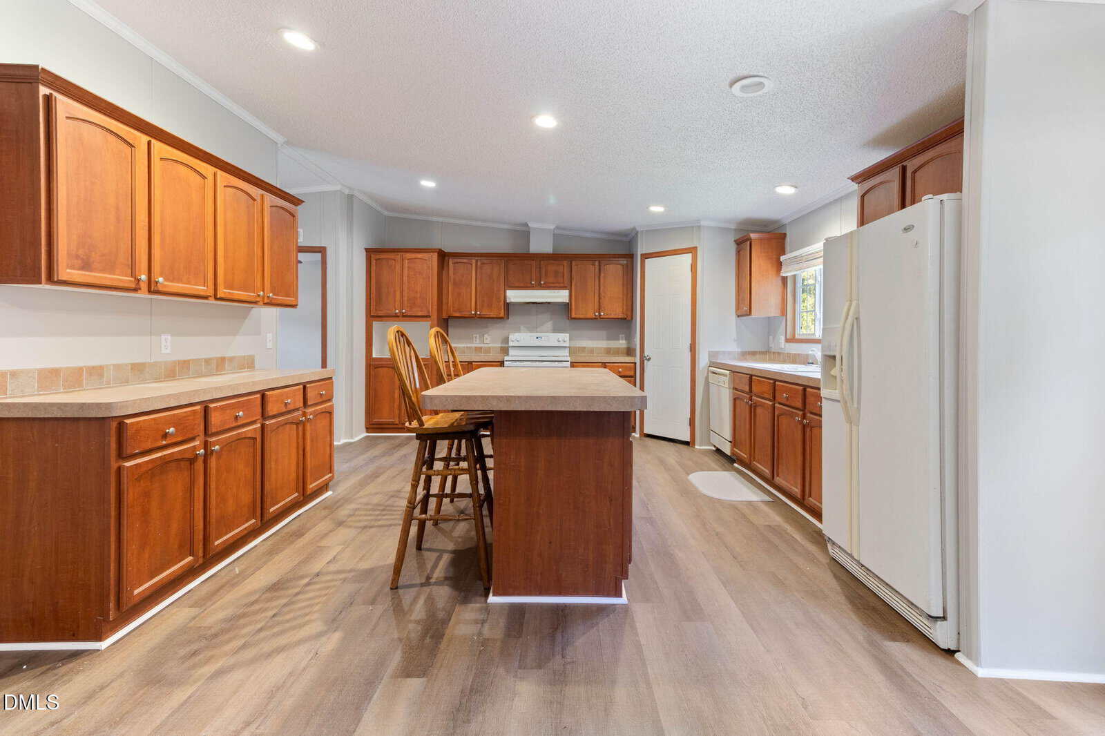 Spacious kitchen with wooden cabinets, white appliances, a center island with two stools, light countertops, laminate wood flooring, and a window above the sink. Dining chairs and a table are seen in the background.