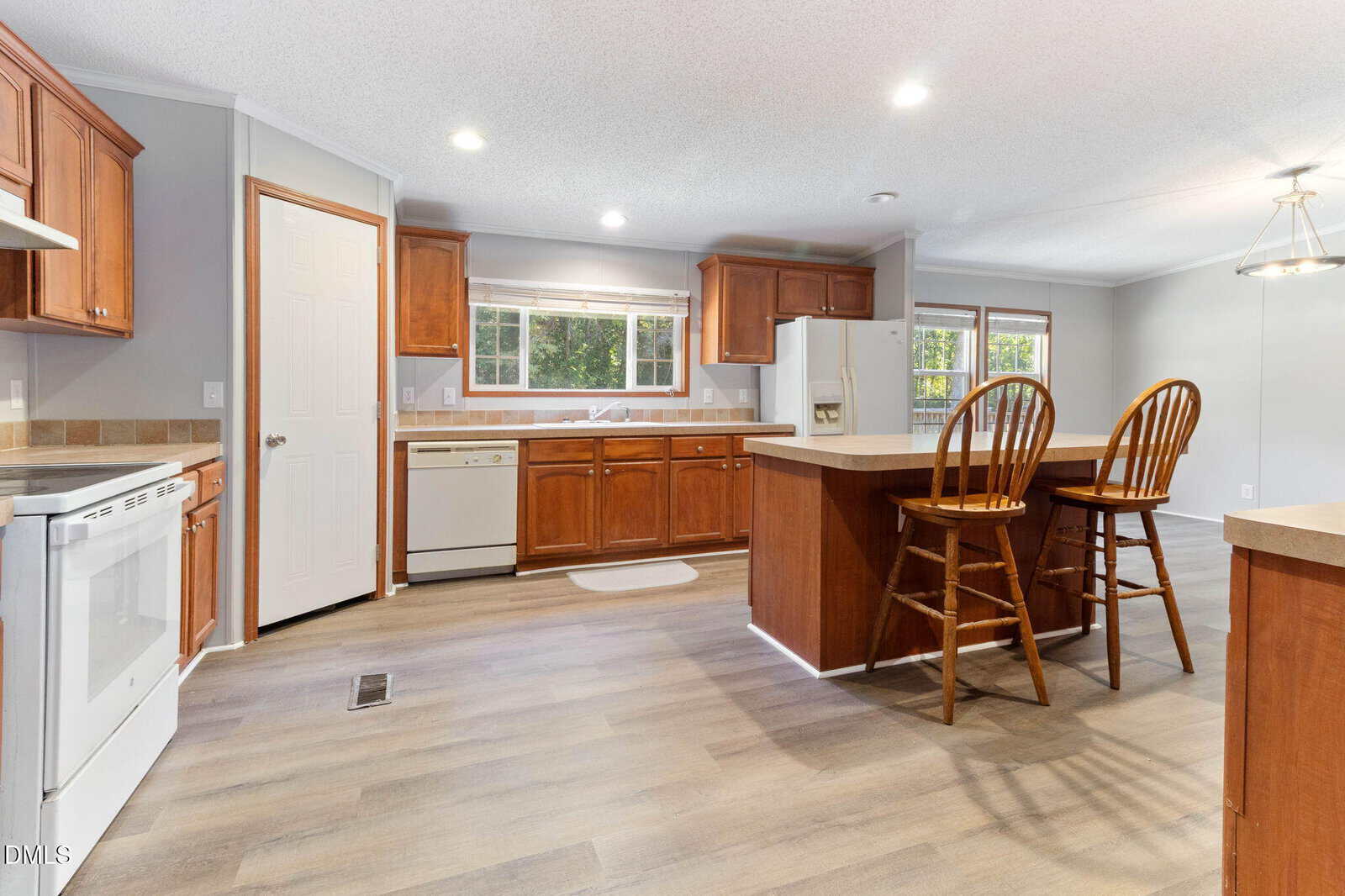 A spacious kitchen with light wood flooring, brown wooden cabinets, white appliances, a central island with two wooden chairs, and large windows letting in natural light.