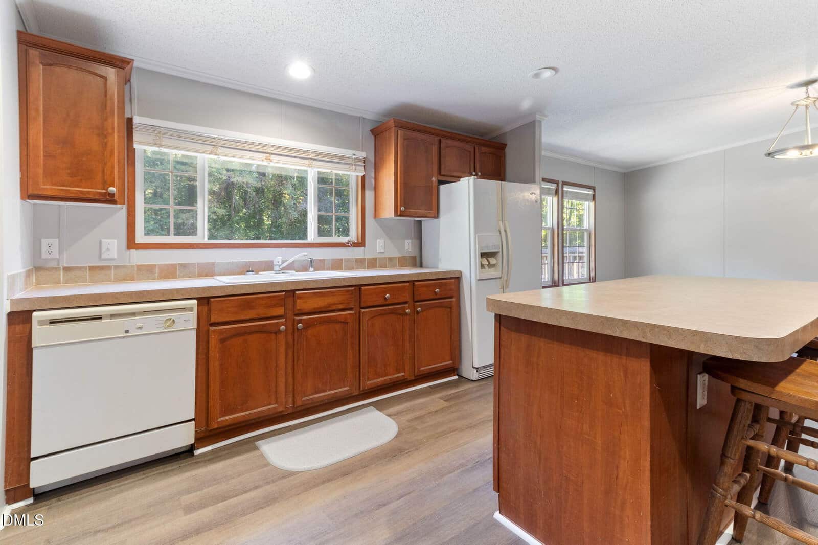 Kitchen with wooden cabinets, white appliances, large window over the sink, and an island with seating. Light wood flooring and neutral walls create a bright, welcoming space.