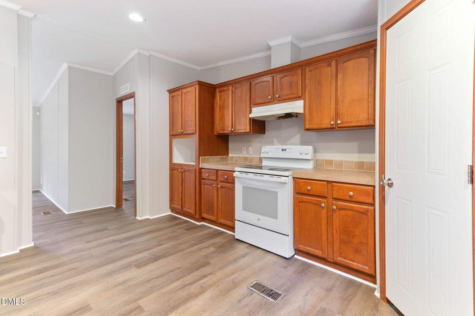 Kitchen with wooden cabinets, white stove and range hood, tan tile backsplash, and light wood flooring. A white door is on the right, and a hallway is visible to the left. Walls are painted light gray.