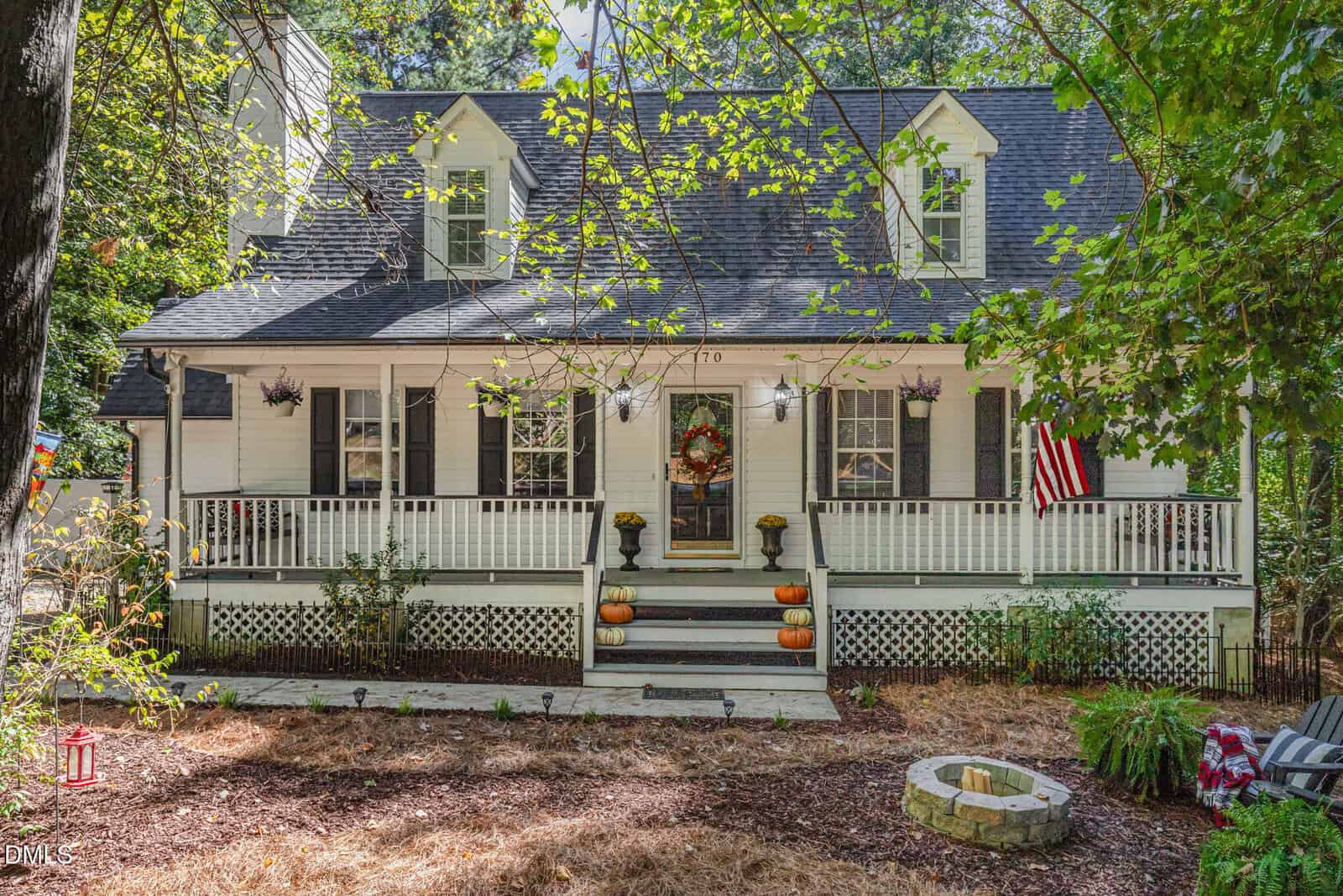 A white house at 170 Misty Way in Franklinton features a front porch, black shutters, and dormer windows, decorated with autumn wreaths and pumpkins. An American flag hangs by the steps, and a fire pit sits in the yard surrounded by trees.