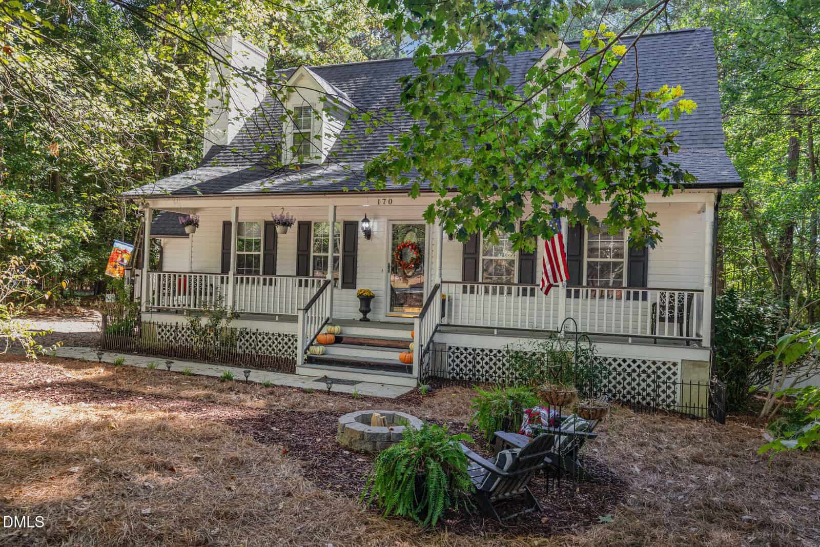 A charming white house with a large front porch decorated for autumn, including pumpkins and a wreath. An American flag hangs near the steps, and outdoor chairs and plants are arranged in the front yard amid surrounding trees.