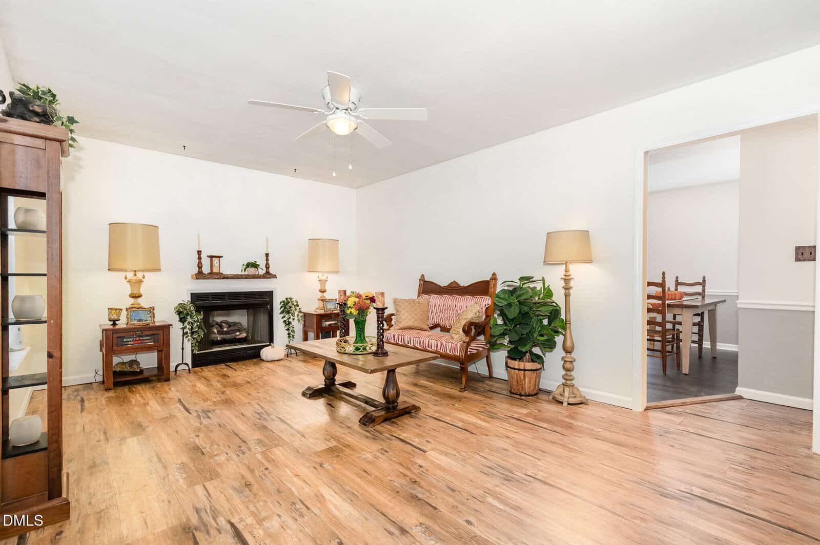 Bright living room with wood flooring, a ceiling fan, fireplace, vintage sofa, coffee table, two tall lamps, plants, and a view into a dining area through an open doorway.