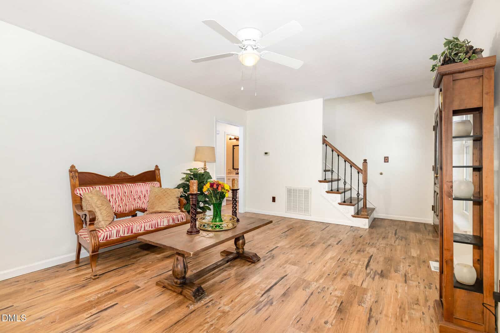 A bright living room with wood flooring, a wooden coffee table, a striped cushioned bench, a ceiling fan, and a staircase with a wooden railing. Shelving with decor items lines one wall.